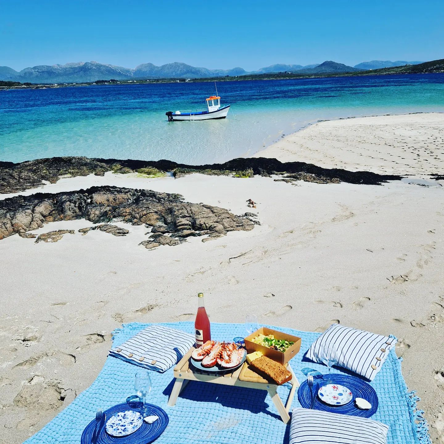 Beach picnic on white sand with a small boat in the turquoise water and the Connemara mountains behind