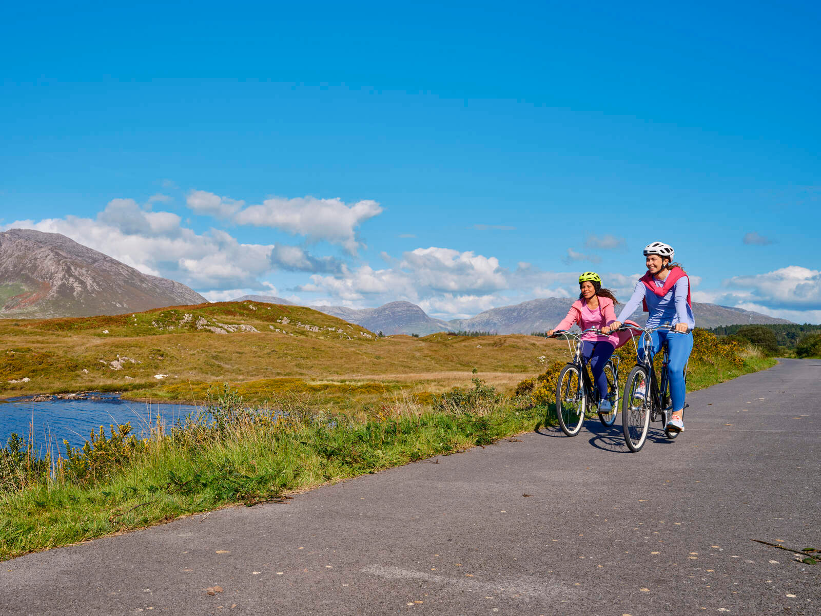 Two women cycling along a quiet Connemara road with mountains and a lake behind