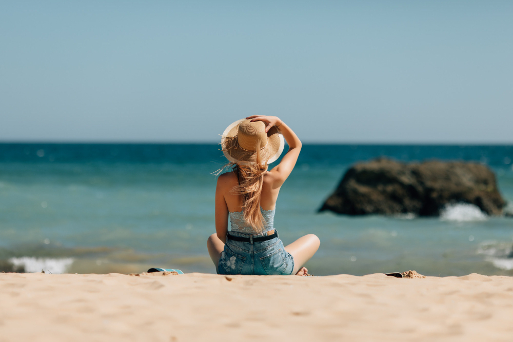 Woman in a sun hat sitting on a sandy beach looking out at the sea
